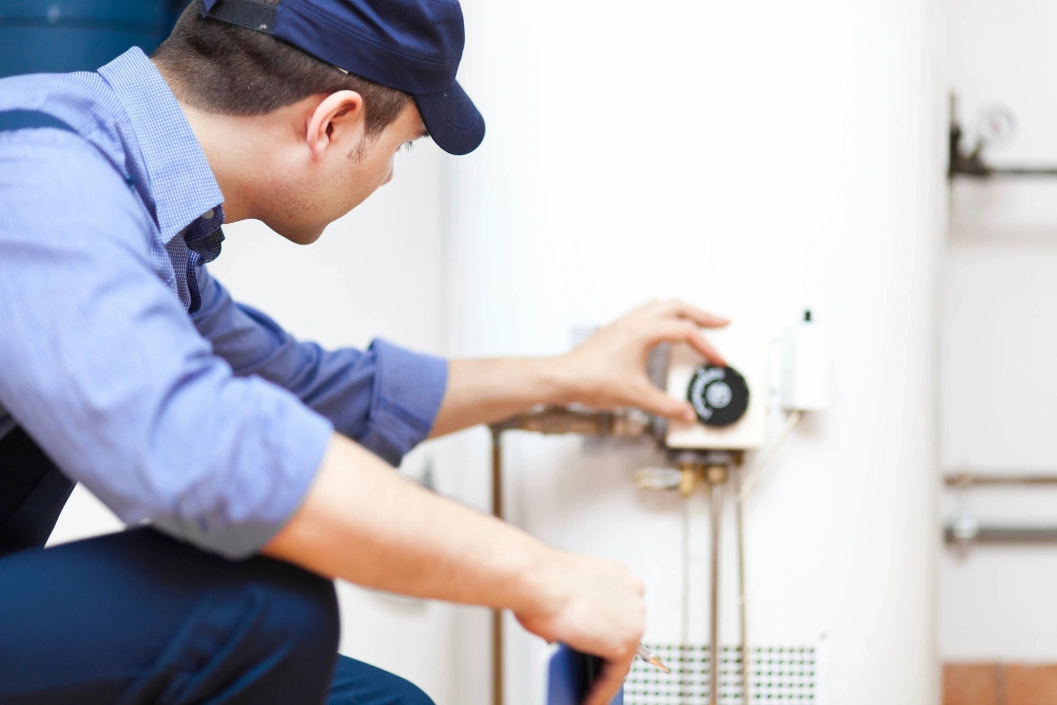 A man in a blue shirt and hat checking a hot water heater.
